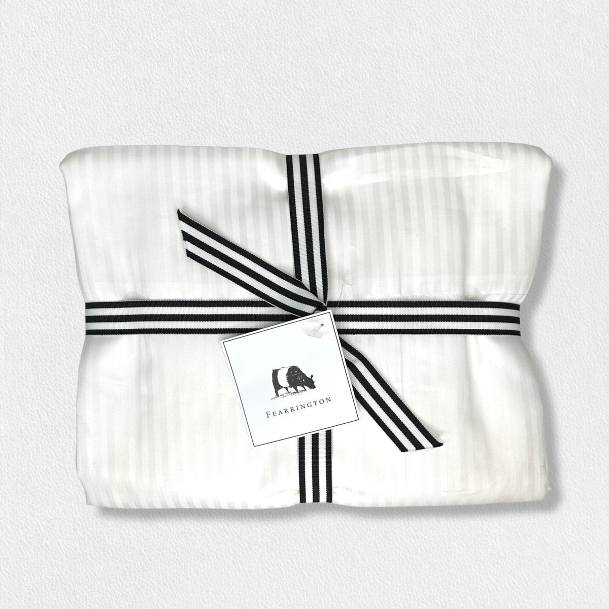 A neatly folded white fabric bundle, tied with a black and white striped ribbon, features a white tag illustrated with a small animal and the text &quot;Fearrington.&quot; The backdrop is an off-white texture, evoking the luxury of the FEARRINGTON LIFESTYLE BEDDING COLLECTION - FRETTE STRIPE KING FLAT SHEET by Frette North America.