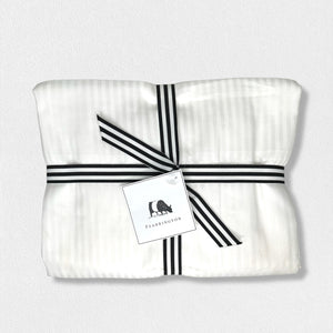 A neatly folded white cloth, secured with a black and white striped ribbon tied into a bow. Attached to the ribbon is a rectangular tag featuring a black silhouette of a cow and the text "FEARRINGTON" below it. Reminiscent of luxury linens, it's set against a plain light gray background. This elegant presentation showcases the FEARRINGTON LIFESTYLE BEDDING COLLECTION - FRETTE QUEEN STRIPE FLAT SHEET by Frette North America.