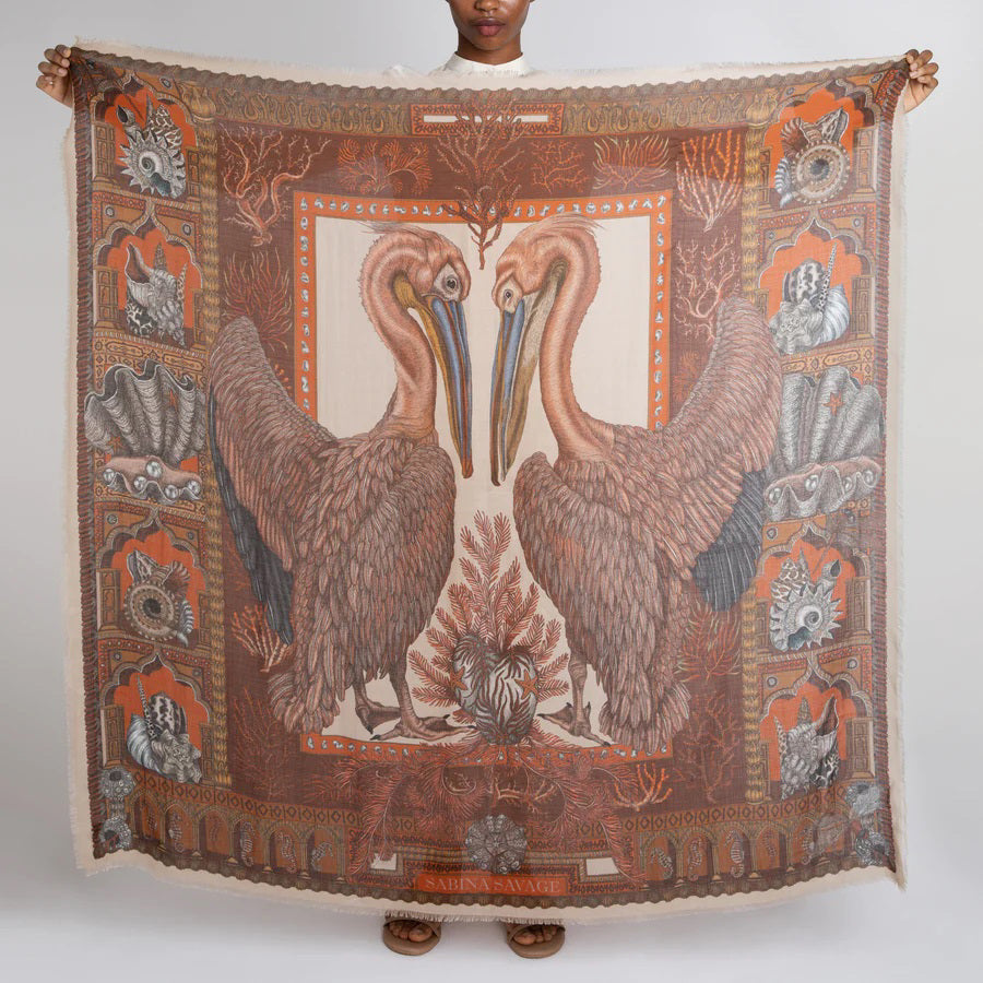 The SABINA SAVAGE - THE PELICANS AND THE SEA IN CASHMERE, VERMILLION/GOLD, 135CM by SABINA SAVAGE ENGLAND is a rectangular scarf adorned with intricate animal and nature-themed patterns in rich brown and orange hues. Made from lightweight cashmere, the scarf showcases a variety of designs including leaves, pink pelicans, and geometric shapes. It is elegantly tied near the top and features fringed edges.