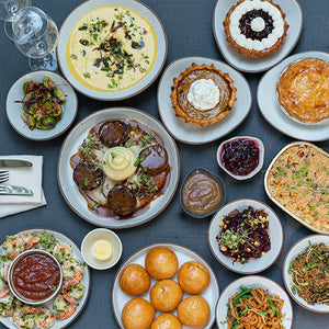 A delightful spread of dishes reminiscent of the Thanksgiving Takeout for Two from FEARRINGTON VILLAGE, available for pickup on November 28, adorns the table. The assortment includes a variety of meats, salads, pies, and rolls. Some plates are embellished with creamy sauces while others boast rich toppings. Glasses and napkins are meticulously arranged on one side.