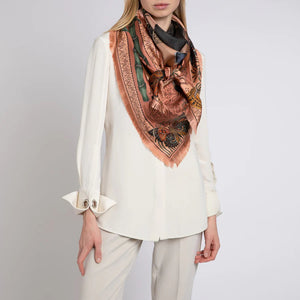 A person wearing a white long-sleeve shirt and white trousers, showcasing the SABINA SAVAGE - THE BUTTERFLY PANDA SILK TWILL 135CM scarf by SABINA SAVAGE ENGLAND. The intricately patterned silk scarf, featuring colors of orange, green, and black, is draped elegantly around their neck like an antique embroidery sampler, while their hands rest by their sides.