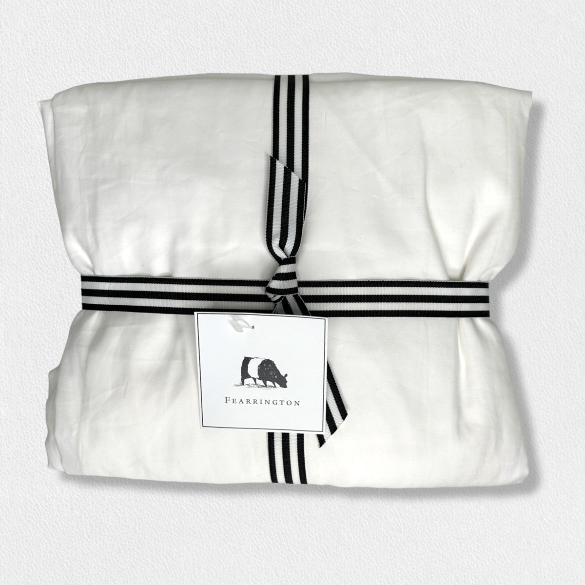 A neatly folded white fabric from the Fearrington Lifestyle Bedding Collection - Frette Queen Fitted Sheets by Frette North America is tied with a black and white striped ribbon. A tag featuring a rooster image and the text &quot;Fearrington&quot; is attached to the ribbon. The background is a plain, light-colored surface, emphasizing the luxury bedding&#39;s elegant presentation.
