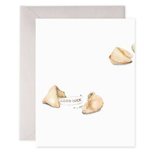 The E. FRANCES - FORTUNE COOKIE CARD, designed by E.FRANCES PAPER, features a minimalistic watercolor painting of a cracked fortune cookie revealing a paper fortune that reads "GOOD LUCK." The card boasts a clean white background printed on luxe heavyweight paper and includes the faint outline of another envelope in the background.