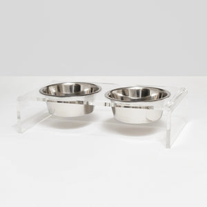 The HIDDEN ELEVATED PET FEEDER 1 QUART (CLEAR) features a clear acrylic stand with four curved legs, elevating two lead-free stainless steel bowls to an ideal height. This minimalist design enhances both style and practicality for your pets, set against a plain light gray surface.