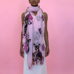 A person stands against a pink background, wearing the SIMONE BRUNS - DOG CASHMERE SCARF adorned with colorful floral and deer prints. The stylish scarf from SIMONE BRUNS is draped around their neck and hangs down to their knees, covering part of their white sleeveless dress.