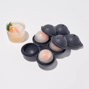 A black SPHERE ICE MOLD by W&P DESIGN, featuring four round compartments with two ice spheres filled, rests on a light gray surface. To the right, a glass holds one of the ice spheres. Two orange halves are positioned in the bottom left corner. This product description emphasizes the sleek and practical design of this 6.3" kitchen accessory.
