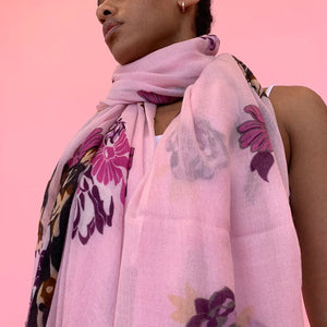 A person stands against a pink background, wearing the SIMONE BRUNS - DOG CASHMERE SCARF adorned with colorful floral and deer prints. The stylish scarf from SIMONE BRUNS is draped around their neck and hangs down to their knees, covering part of their white sleeveless dress.