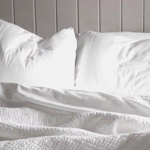 A neatly folded white cloth, secured with a black and white striped ribbon tied into a bow. Attached to the ribbon is a rectangular tag featuring a black silhouette of a cow and the text "FEARRINGTON" below it. Reminiscent of luxury linens, it's set against a plain light gray background. This elegant presentation showcases the FEARRINGTON LIFESTYLE BEDDING COLLECTION - FRETTE QUEEN STRIPE FLAT SHEET by Frette North America.