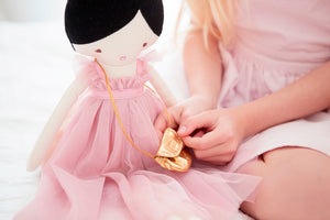 The ALIMROSE - CHARLOTTE DOLL BLUSH by ALIMROSE is a charming doll with black hair styled in a bun, dressed in a pink tulle dress with a high neckline and carrying a gold purse over her shoulder. Suitable for children aged 3 and up, the doll features simple embroidered facial details and a small, painted heart-shaped mouth.