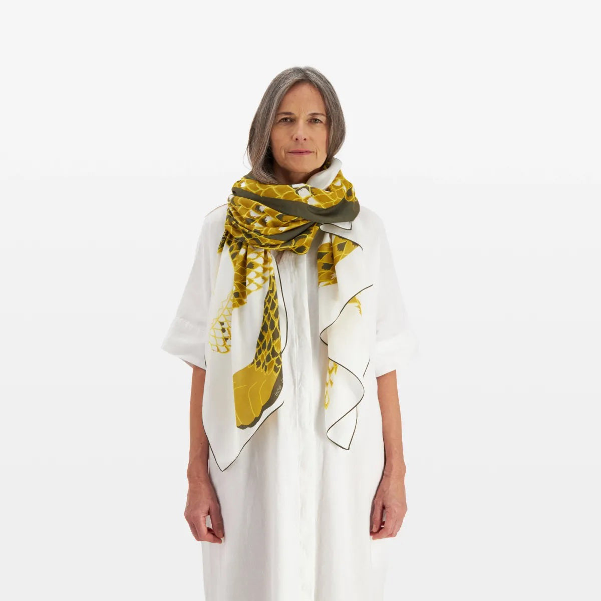 A woman with long, grey hair is wearing a white, loose-fitting dress and has an INOUI EDITIONS - NEOFELIS cotton and silk scarf from INOUI, featuring a snake pattern in yellow, draped around her shoulders. She stands against a plain, white background, looking directly at the camera.