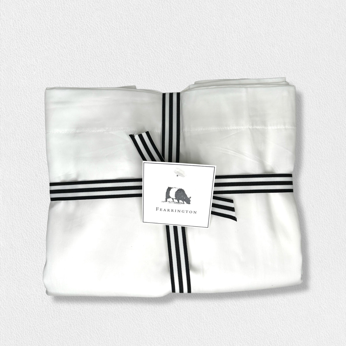 A neatly folded set of white cotton sateen pillowcases from the Fearrington Lifestyle Bedding Collection by Frette North America, boasting a luxurious 300 thread count, is tied with a black-and-white striped ribbon. Attached to the ribbon is a label featuring a small black silhouette of a person seated next to a large bear and the text &quot;Fearrington.&quot; The background is light gray.