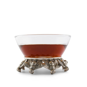 The HONEYBEE GLASS BOWL by VAGABOND HOUSE rests on an ornate stand adorned with intricately detailed wings of pure-pewter bees. The metallic base features multiple bees in a symmetrical arrangement, providing a decorative and elegant support for the bowl above.