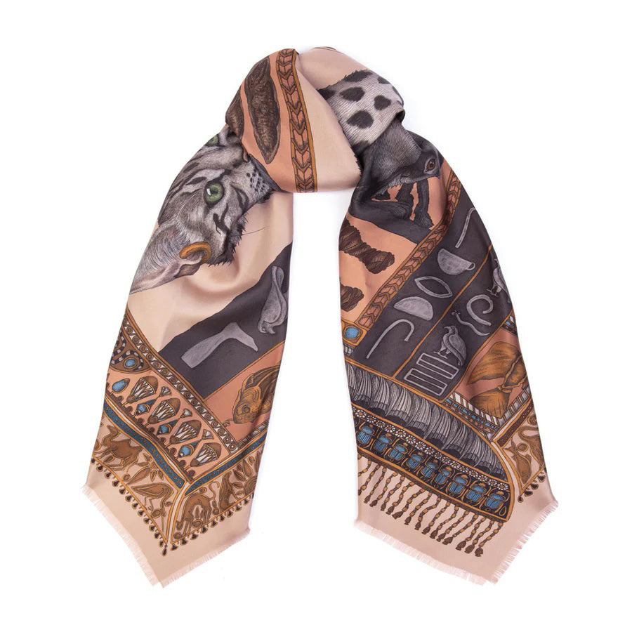 The SABINA SAVAGE - THE BASTET'S BOUNTY IN SILK TWILL 135CM by SABINA SAVAGE ENGLAND is a rectangular scarf made from 100% silk. It features an exquisite design adorned with animal motifs, including a tiger and an Egyptian Mau cat. The edges showcase intricate patterns in shades of brown, orange, blue, and gold. Draped in a simple wrap style, it allows the elaborate artwork to take center stage. Truly Bastet's Bounty.
