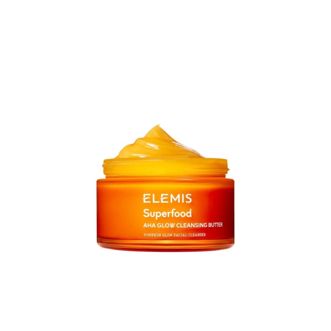 A bright orange jar with a white lid, labeled &quot;ELEMIS - SUPERFOOD AHA GLOW CLEANSING BUTTER&quot; in white text, from the brand ELEMIS - STEINER, featuring nourishing Mango Butter and Pumpkin enzymes. The jar is centered against a plain white background.