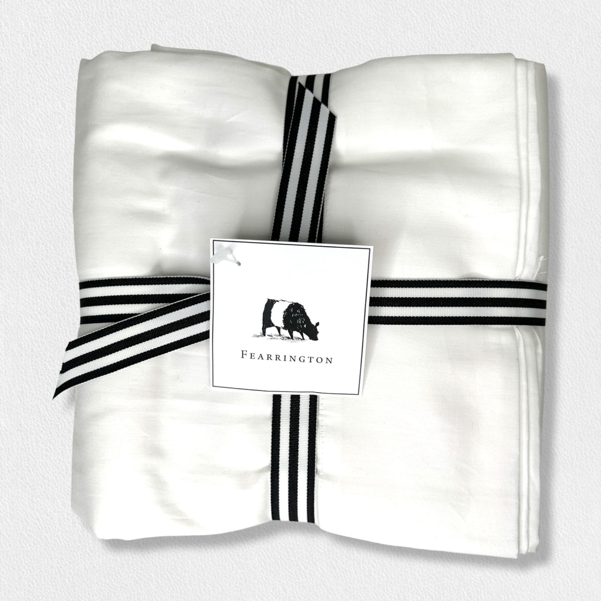 A neatly folded white fabric from the Fearrington Lifestyle Bedding Collection - Frette Queen Flat Sheet is tied with a black and white striped ribbon. A tag attached to the ribbon features a small black silhouette of a cow with &quot;Fearrington&quot; below it, hinting at the luxury bedding&#39;s refined elegance. The background is plain white.