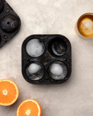 A black SPHERE ICE MOLD by W&P DESIGN, featuring four round compartments with two ice spheres filled, rests on a light gray surface. To the right, a glass holds one of the ice spheres. Two orange halves are positioned in the bottom left corner. This product description emphasizes the sleek and practical design of this 6.3" kitchen accessory.
