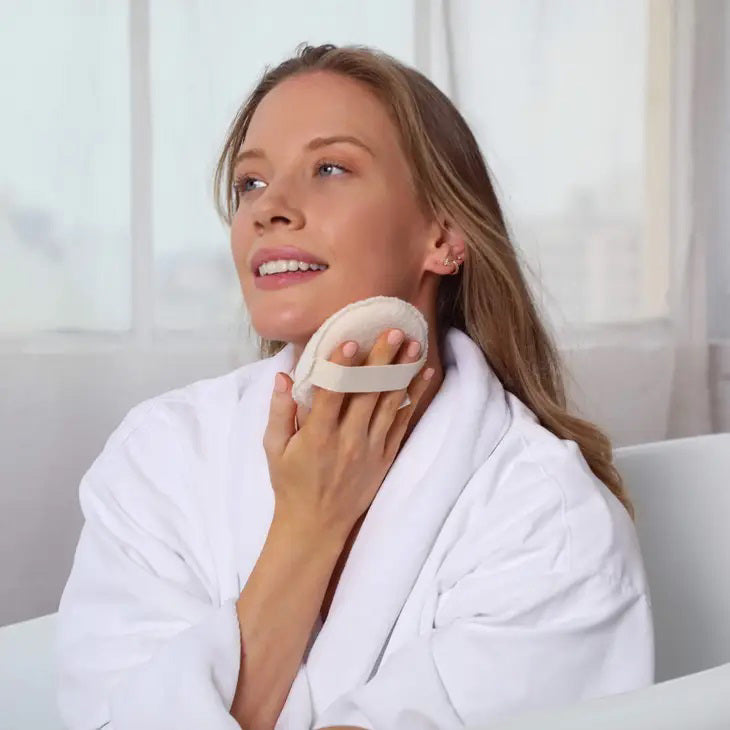 The DAILY CONCEPTS EXFOLIATING BODY SCRUBBER, enclosed in a clear plastic bag with branding details, features a circular design with dual textures for the ultimate exfoliation experience. One side is soft while the other is more abrasive. A beige strap on the back ensures easy grip and enhances your skin’s glow.