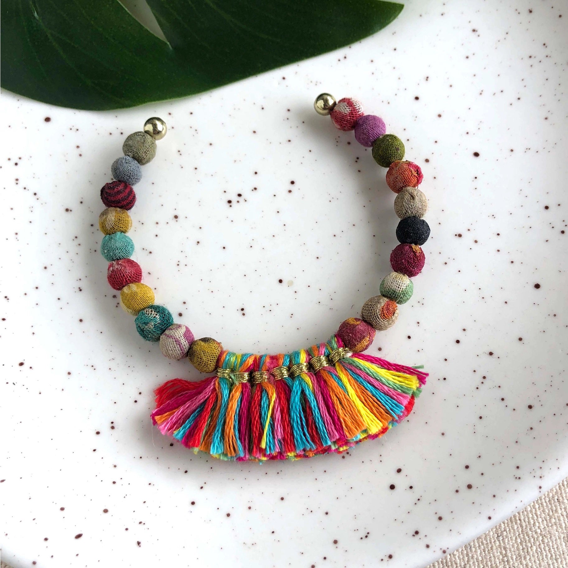 A colorful beaded bracelet lies on a white speckled plate. The KANTHA FRINGE CUFF from WORLD FINDS features small round beads in various bright colors and a vibrant tassel made of multicolored threads, forming a semicircle pattern. A green leaf partially overlaps the plate in the background, enhancing its boho look.