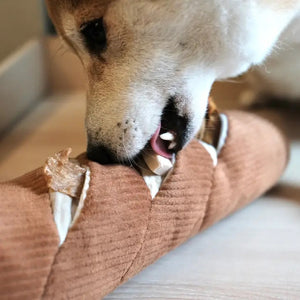 The LAMBWOLF COLLECTIVE - BAGUETTE DOG TOY, by LAMBWOLF COLLECTIVE, resembling a French baguette loaf, rests atop a white cloth. This oversized pet toy showcases light brown textured fabric with cream-colored slits representing the scoring marks typical of baked bread.