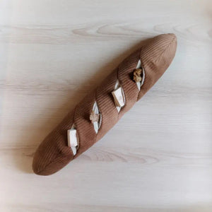 The LAMBWOLF COLLECTIVE - BAGUETTE DOG TOY, by LAMBWOLF COLLECTIVE, resembling a French baguette loaf, rests atop a white cloth. This oversized pet toy showcases light brown textured fabric with cream-colored slits representing the scoring marks typical of baked bread.