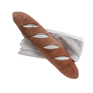 The LAMBWOLF COLLECTIVE - BAGUETTE DOG TOY, by LAMBWOLF COLLECTIVE, resembling a French baguette loaf, rests atop a white cloth. This oversized pet toy showcases light brown textured fabric with cream-colored slits representing the scoring marks typical of baked bread.