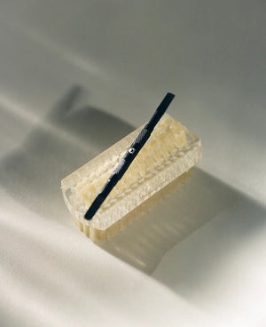 A black TENOVERTEN - THE PUSH BACK CUTICLE tool with a textured handle rests diagonally atop a rectangular, translucent soap bar. The background is a light, neutral shade, and soft shadows are cast from the objects. This minimal and clean image emphasizes salon-grade precision in grooming essentials by TEN OVER TEN.