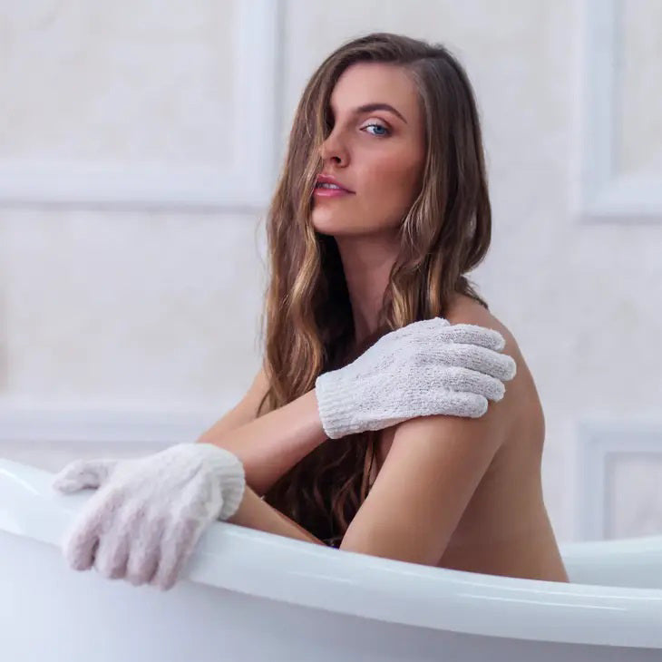 Packaging of DAILY CONCEPTS EXFOLIATING GLOVES next to one pair of white exfoliating gloves. The packaging features a purple and white design with product information, including &quot;Individuals who think in the shower,&quot; an &quot;organic cotton&quot; icon, and tips for use as a Pre Self-Tanning Ritual.